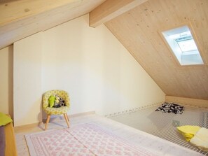 Una habitación con techo inclinado, un ventanal pequeño, una silla con juguetes, un tapete estampado y una cama con colchón a rayas.