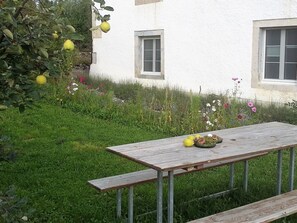 A wooden table with fruits and a bench in a garden with a white building in the background.