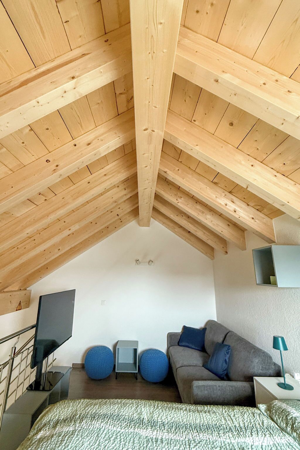 Ein Schlafzimmer im Loft-Stil mit freiliegenden Holzbalken, einem grauen Sofa, zwei blauen runden Hocker, einem kleinen Fernseher und einer Nachttischlampe.