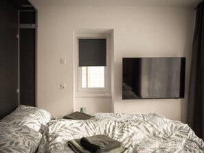 Ein Hotelzimmer mit Bett, einem Fenster mit Jalousie, einem an der Wand befestigten Flachbildfernseher und einer Tür zu einem anderen Raum.