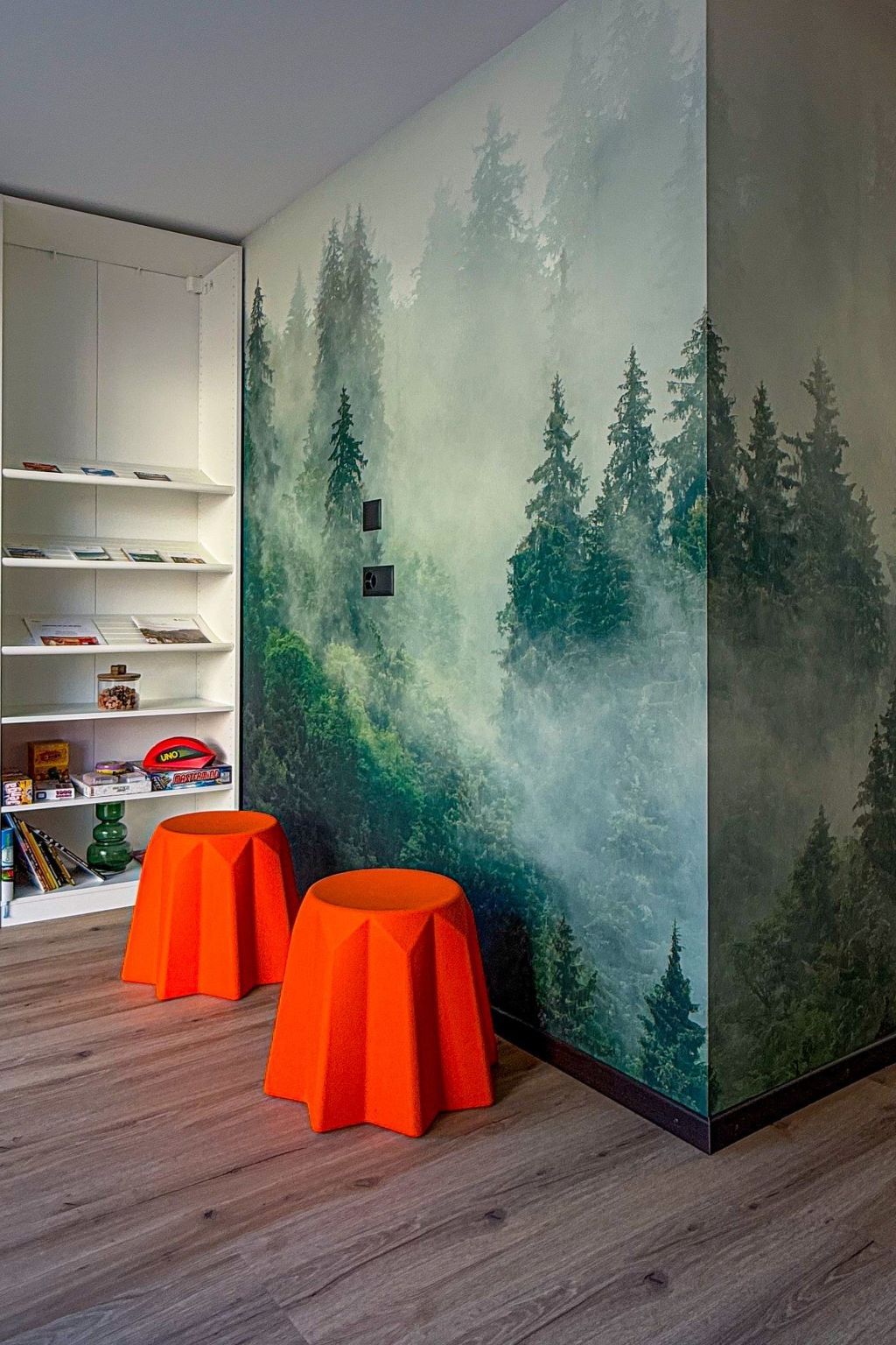 Een kamer met een boekenkast, twee oranje krukken en een bosdecoratie op de muur.
