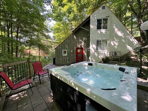 A hot tub on a patio with a wooden deck, a red chair, and a small shed in a wooded area. - Chalets Cheribourg - Le 144 - Spa (Orford)