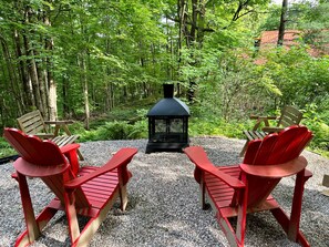 A forested area with two red Adirondack chairs facing a black lantern-style fire pit. - Chalets Cheribourg - Le 144 - Spa (Orford)
