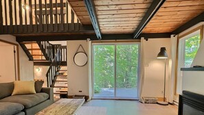 A modern living room with a sofa, a wooden coffee table, a staircase, and a large sliding glass door leading to an outdoor area. - Chalets Cheribourg - Le 144 - Spa (Orford)