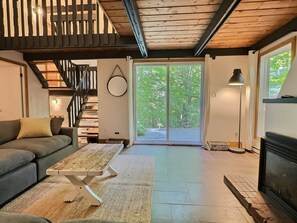 A modern living room with a sofa, a wooden coffee table, a staircase, and a large sliding glass door leading to an outdoor area. - Chalets Cheribourg - Le 144 - Spa (Orford)