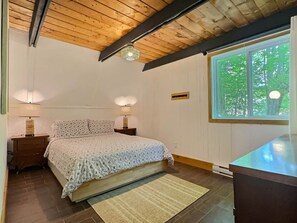 Ein Schlafzimmer mit Holzboden, einem Bett mit gemustertem Überwurf, zwei Nachttischlampen, einem Fenster mit Blick auf Bäume und einer kleinen Schreibtischzeile.