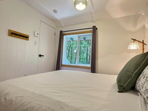 A bedroom with a bed, a window with curtains, a wall-mounted lamp, and a door.