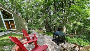 A patio with Adirondack chairs, a wooden table, and a black metal fire pit surrounded by gravel and greenery. - Chalets Cheribourg - Le 144 - Spa (Orford)