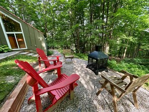 A patio with Adirondack chairs, a wooden table, and a black metal fire pit surrounded by gravel and greenery. - Chalets Cheribourg - Le 144 - Spa (Orford)