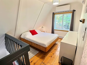 Un dormitorio con cama de madera, almohada roja, lámpara de noche, ventana con vistas a árboles y unidad de aire acondicionado blanca.