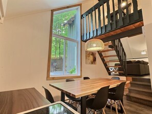 A dining area with a wooden table and chairs, a large window with a view of trees, a staircase with a black railing, and a hanging light fixture.