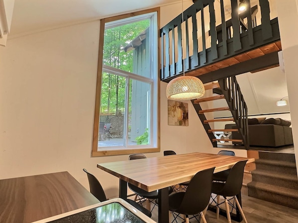 Ein Essbereich mit Holztisch und -stühlen, ein großes Fenster mit Blick auf Bäume, eine Treppe mit schwarzer Geländer und einer hängenden Leuchte.