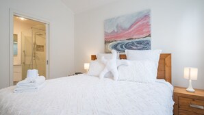 Una cama bien tendida con sábanas y almohadones blancos, un cabecero de madera, un cuadro en la pared y una mesita de noche con una lámpara.