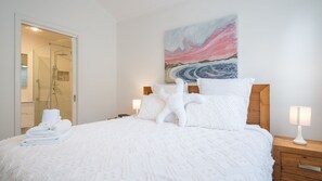 Ein ordentlich bezogenes Bett mit weißen Leinen und Kissen, ein Holzkopfstück, ein Gemälde an der Wand und ein Nachttisch mit Lampe.
