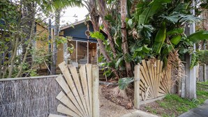 Una reja de madera con diseño de abanico, una casa con ventana visible y abundante vegetación tropical.