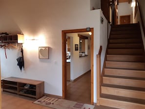 Een trap naar de tweede verdieping, een houten bankje, een kapstok aan de muur en een keuken in de achtergrond.