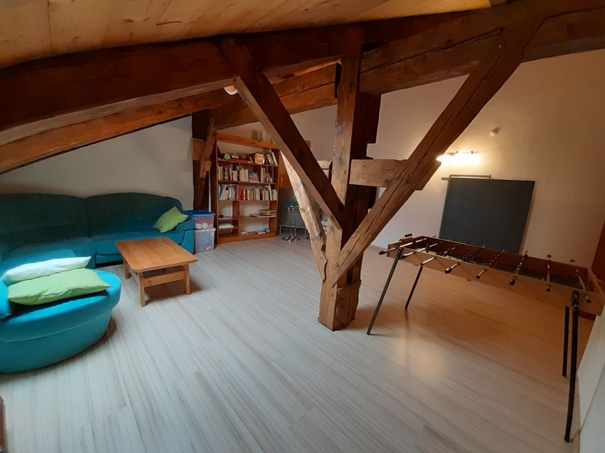 A loft-style room with a wooden ceiling, a blue sofa, a small wooden table, a bookshelf, and a foosball table.