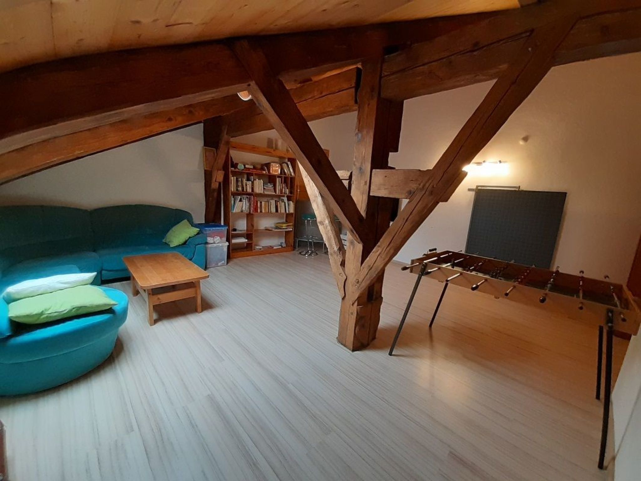 A loft-style room with a wooden ceiling, a blue sofa, a small table, a bookshelf, and a foosball table.