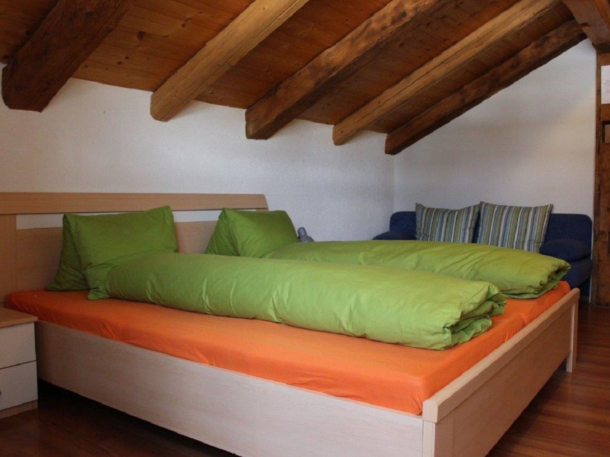 A bedroom with a wooden ceiling, a bed with green and orange bedding, and a blue sofa with striped cushions.