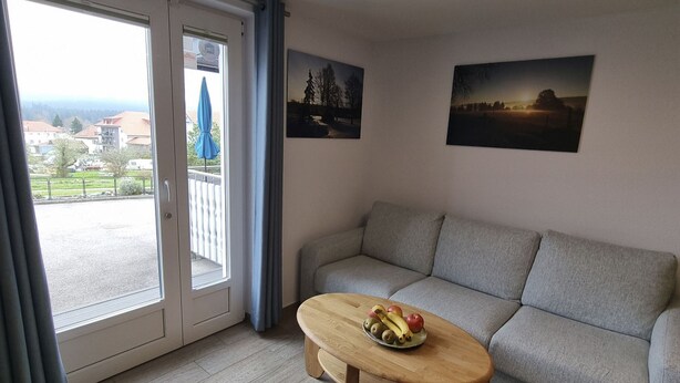 Ein Wohnzimmer mit grauem Sofa, einem Couchtisch mit Früchten, einem Balkon mit blauem Sonnenschirm und zwei Landschaftsbildern an der Wand.
