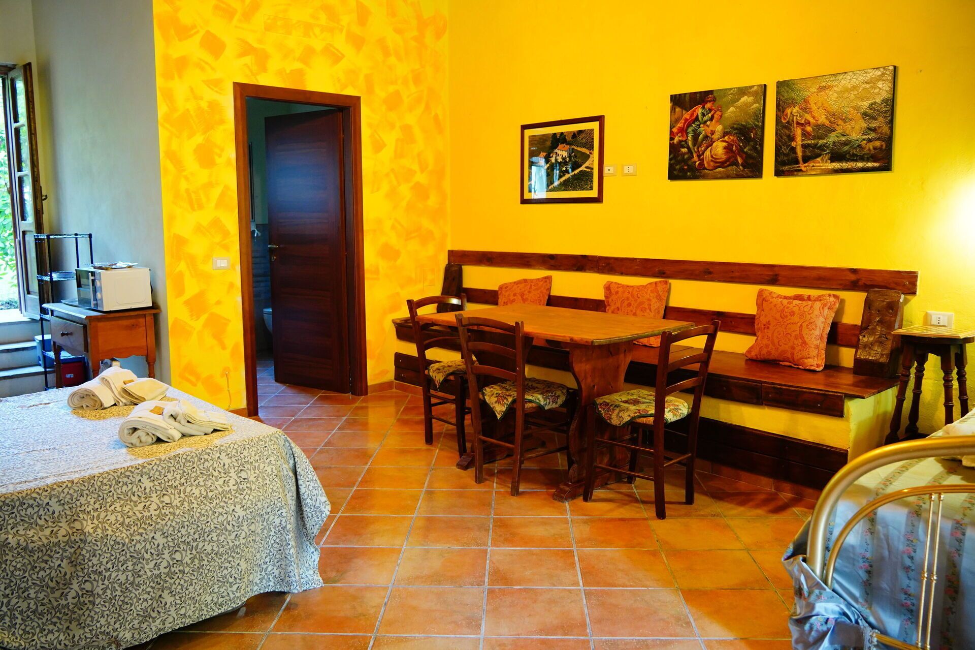 Una habitación con una cama, una mesa de comedor de madera con sillas, un banco y dos cuadros en la pared.