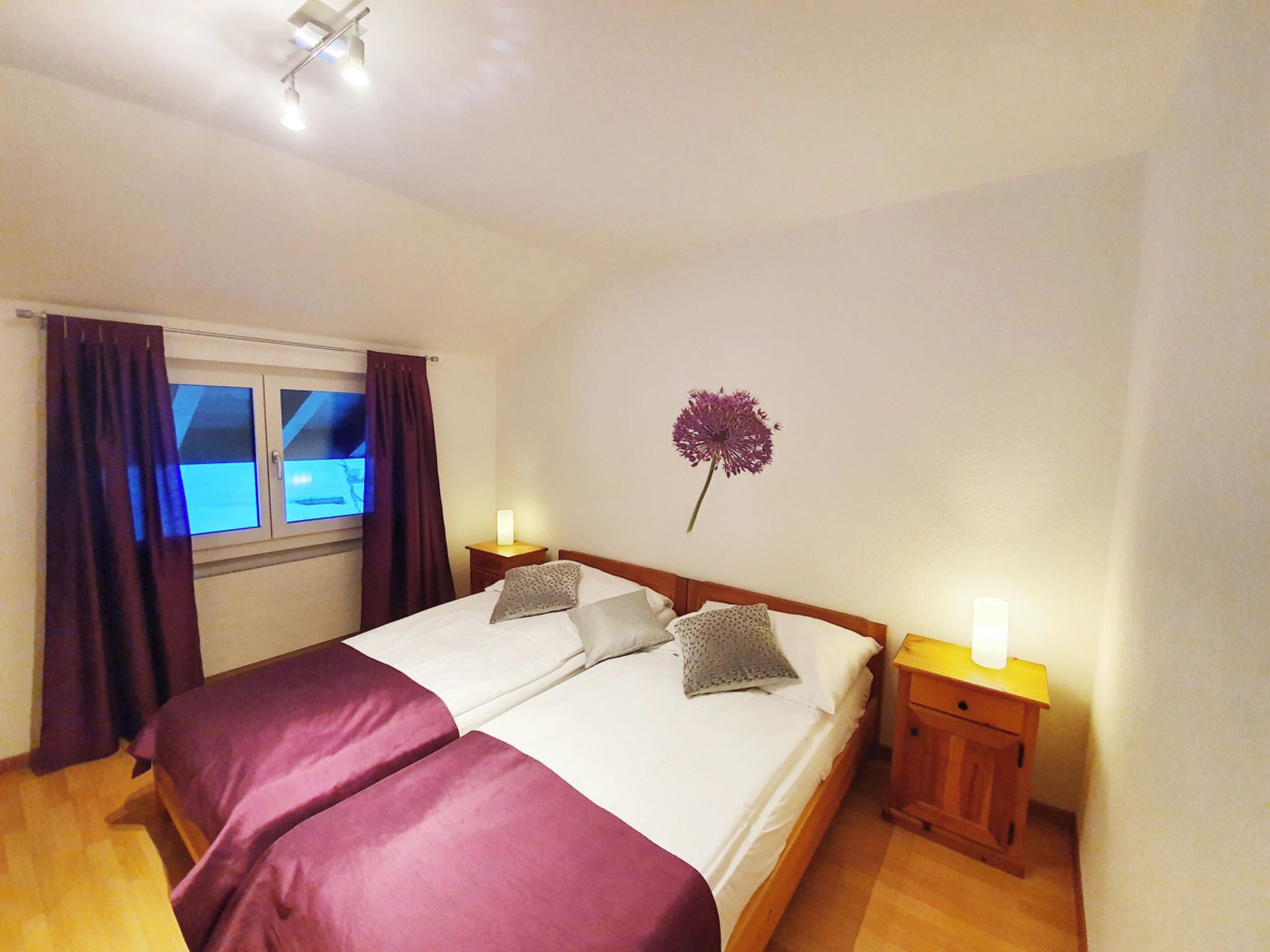 Un lit double avec deux oreillers, une table de chevet avec une lampe, une fenêtre avec des rideaux et une plante en pot fixée au mur.