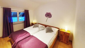 Una cama doble con dos almohadas, una mesita de noche con lámpara, una ventana con cortinas y una flor en la pared.