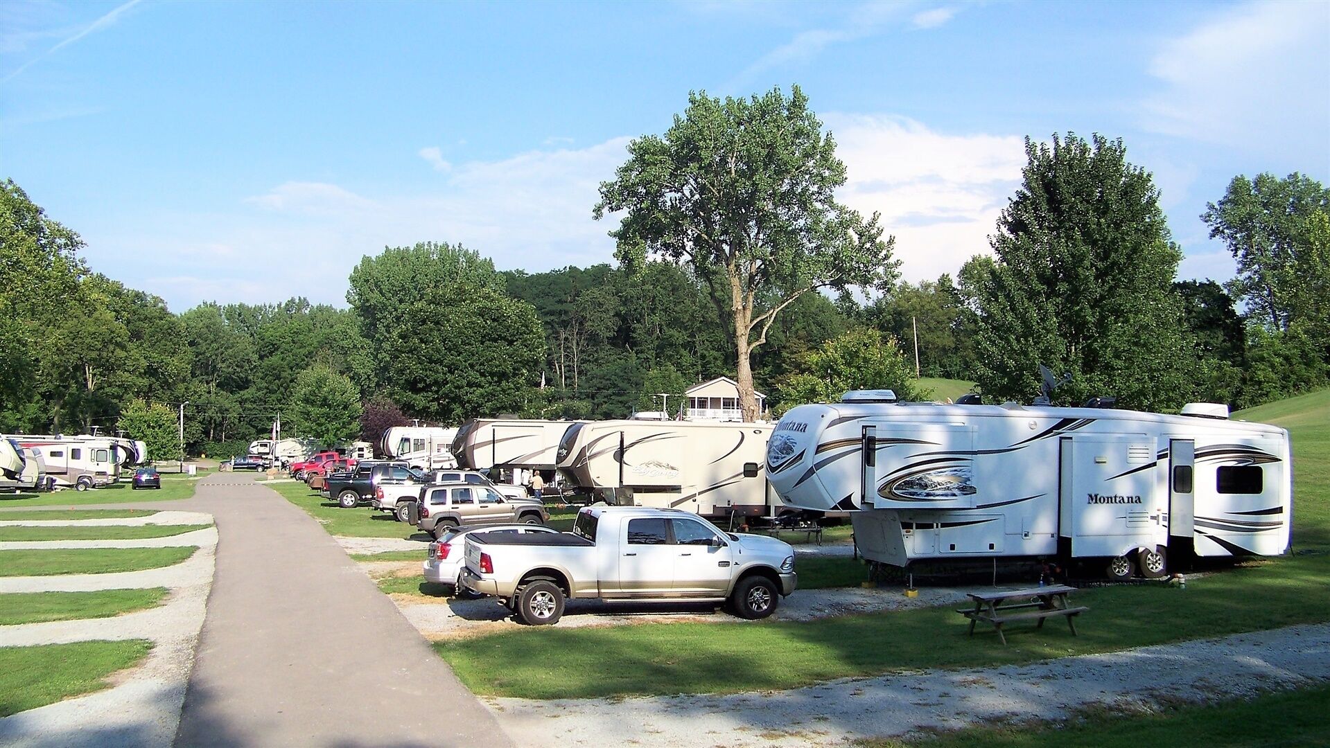 Un terrain de camping où l’on peut voir des camping-cars et des camions stationnés sur des pelouses, un chemin pavé et des arbres en arrière-plan.