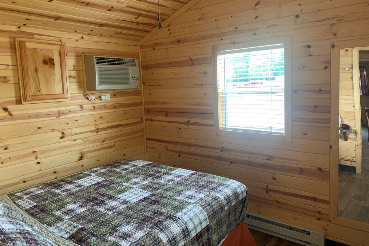 Una habitación de cabaña de madera con una cama, un equipo de aire acondicionado, una ventana y una puerta.