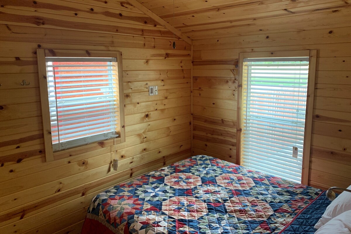 Una habitación de cabaña de madera con una cama, dos ventanas con persianas, un ventilador de techo y una lámpara fijada a la pared.
