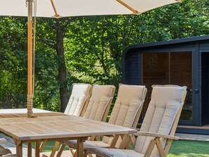 Un espace extérieur aménagé avec une table et des chaises en bois, placé sous une tonnelle et entouré de verdure.