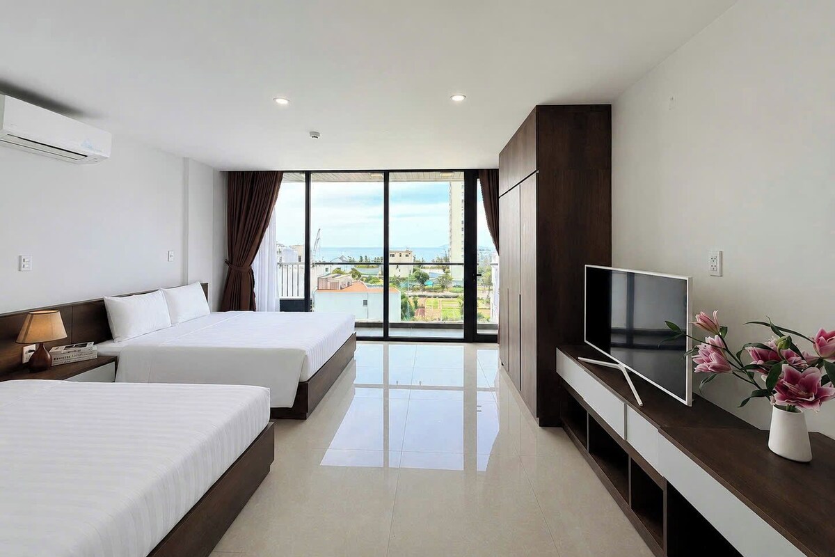 A hotel room with two beds, a TV, a desk, and a large window with a city view.