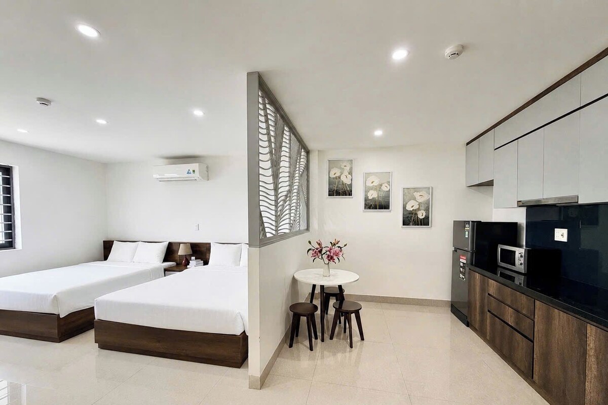 A modern hotel room with two beds, a small dining table, and a kitchenette.