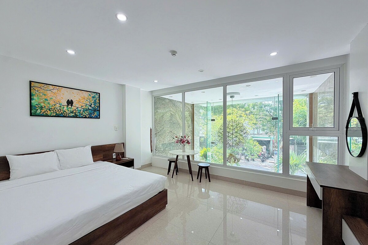 A modern bedroom with a large bed, a dining table with chairs, and a view of greenery through large windows.