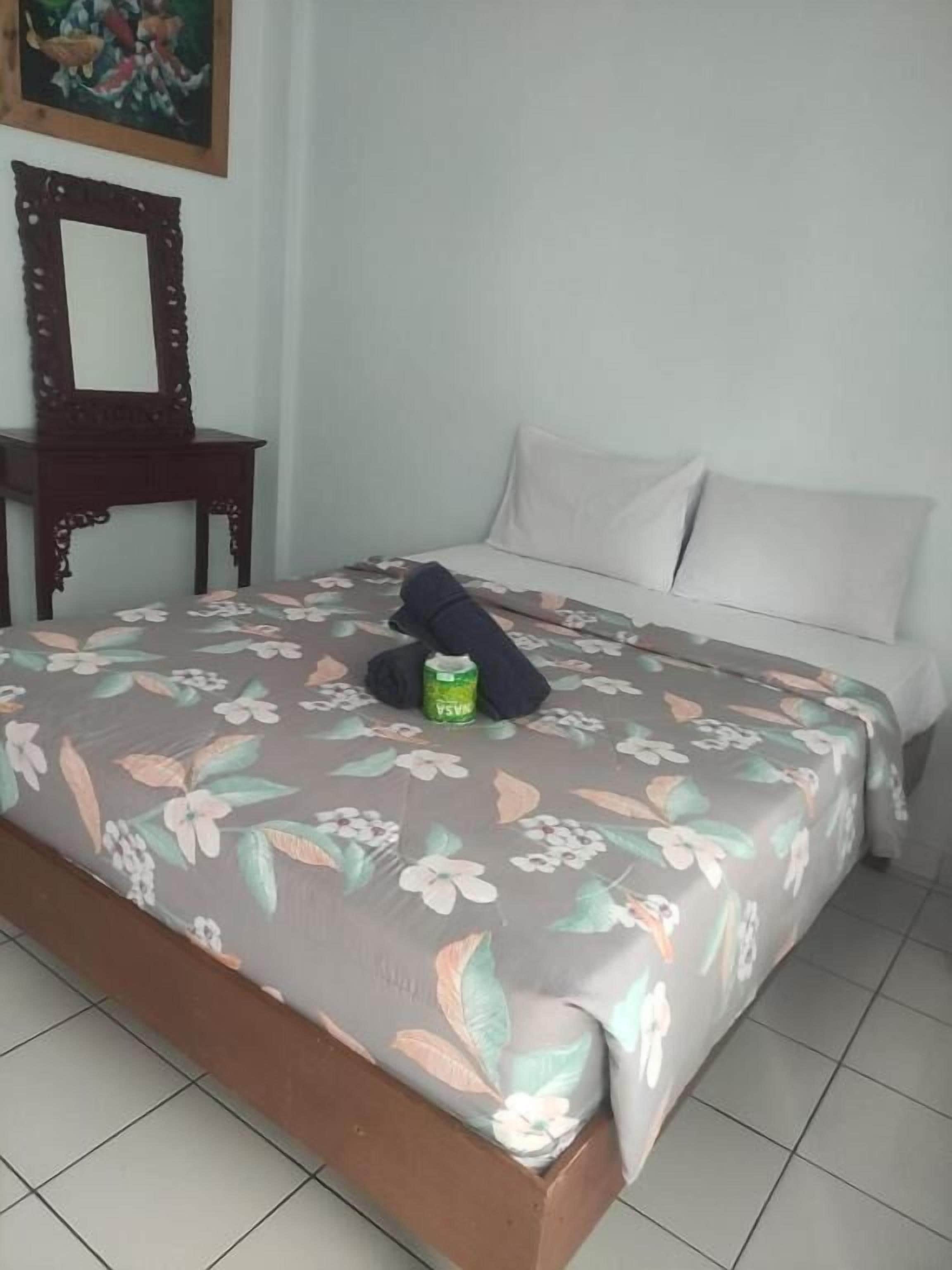 Una cama con cubrecama floral, una mesita de noche de madera con espejo y almohadas blancas.
