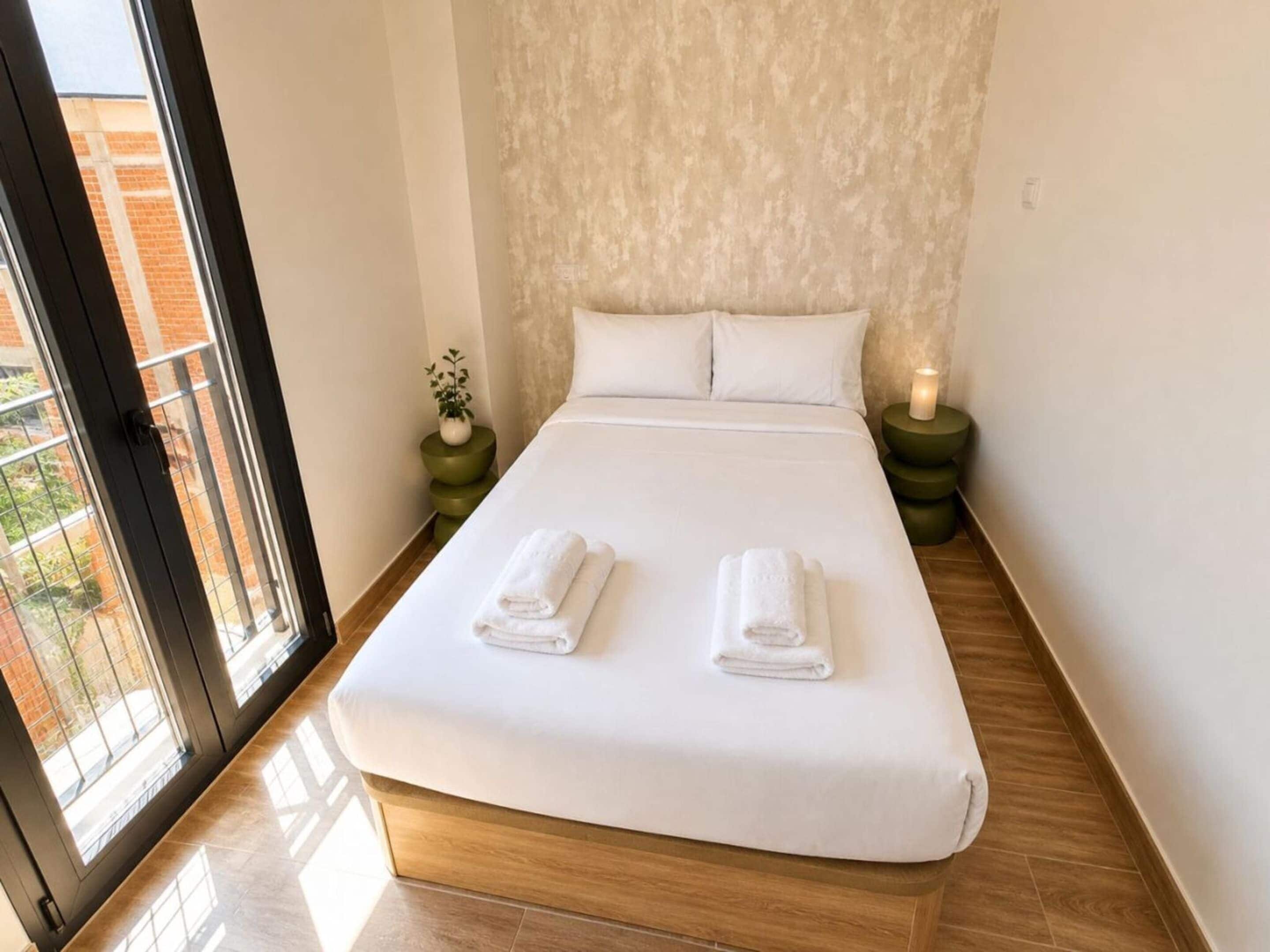 Habitación de hotel con una cama grande, dos almohadas, una mesita de noche con una vela, y vista a un edificio de ladrillo a través de una puerta de vidrio.