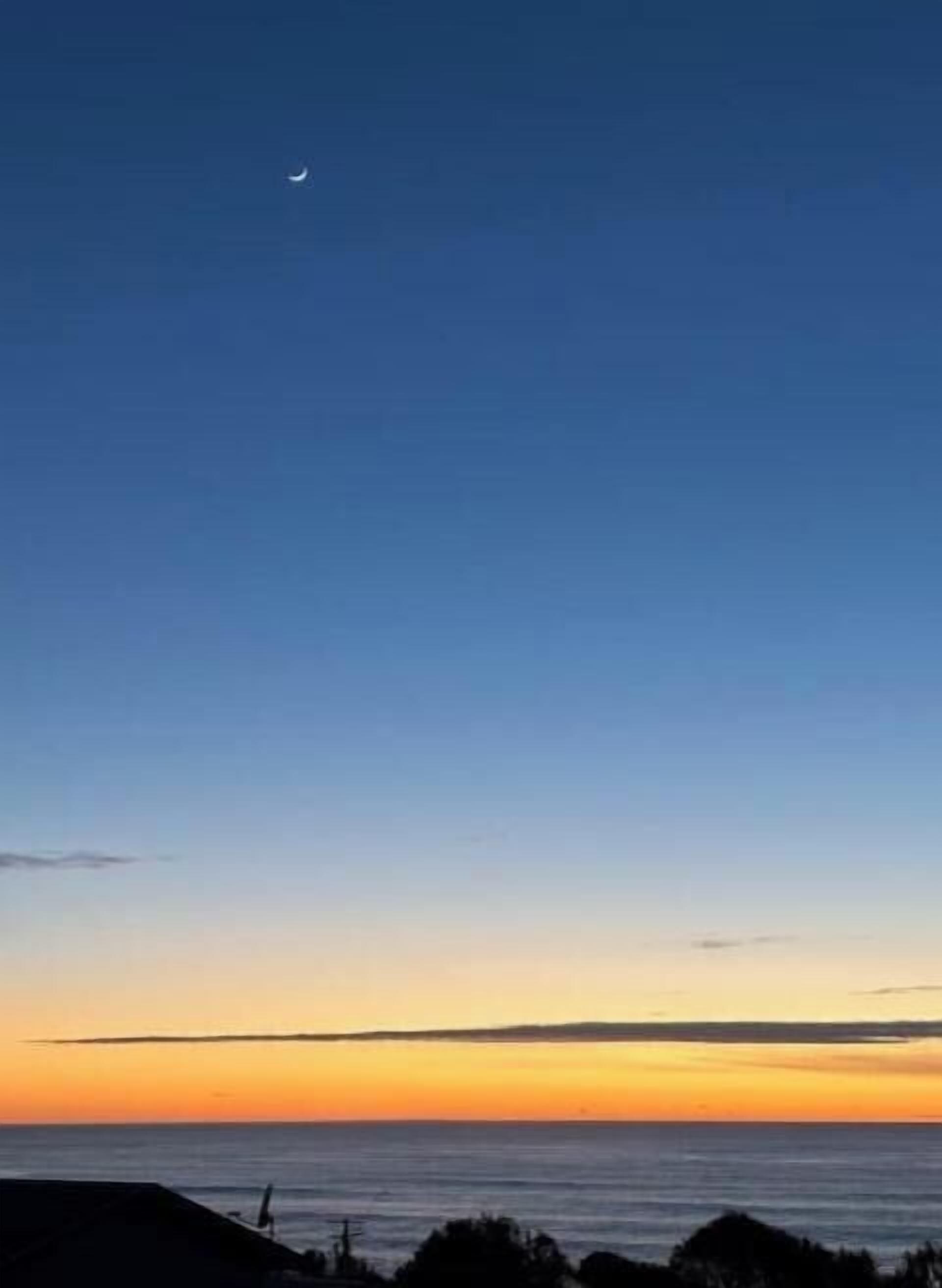 A coastal sunset with a crescent moon, calm sea, and silhouetted land.
