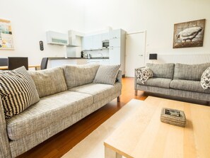 A living room with a sofa and a coffee table, a kitchen in the background, and a painting on the wall.