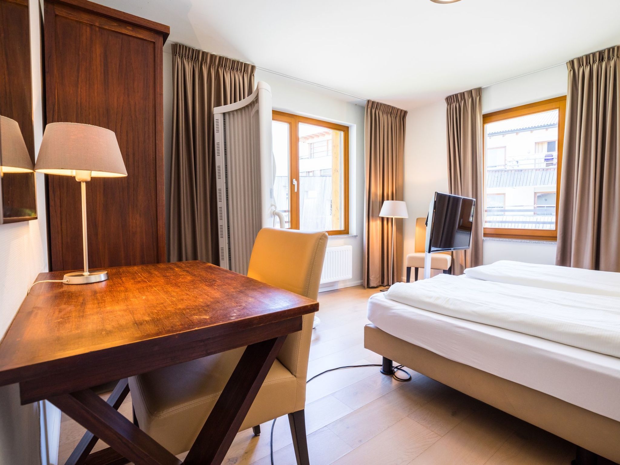 Una habitación de hotel con un escritorio de madera, una cama, un televisor y ventanas grandes con cortinas.