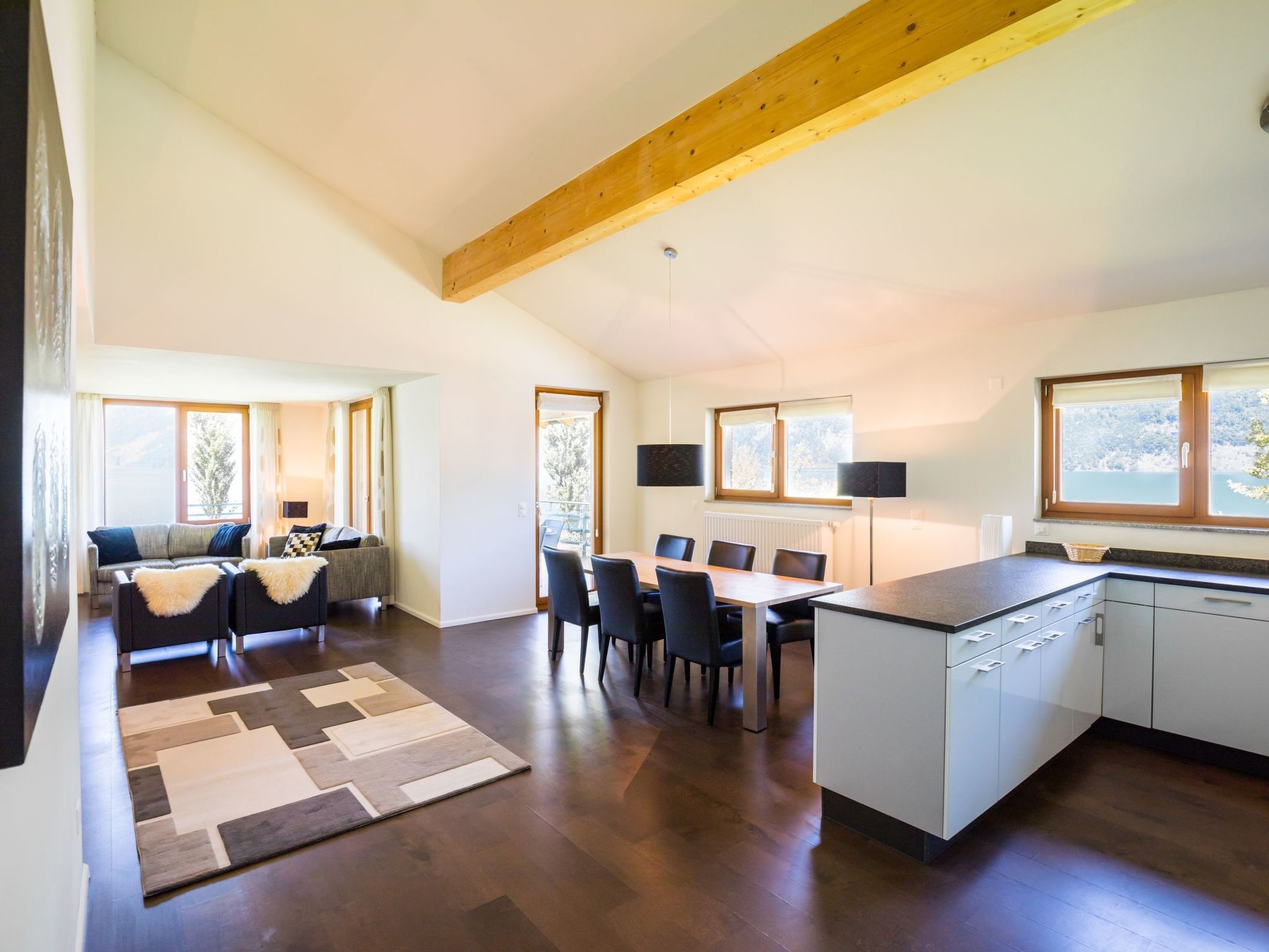 A modern kitchen with a dining area, wooden ceiling beams, and large windows.