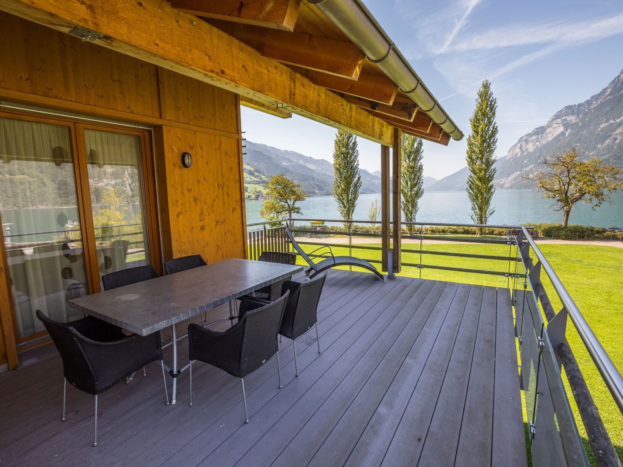 A wooden deck with a table and chairs, overlooking a lake and mountains.