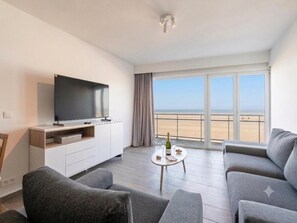 A modern living room with a flat-screen TV, grey sofas, a small coffee table with a bottle and glasses, and a large window with a beach view. - Classy studio with sea view. (Blankenberge)