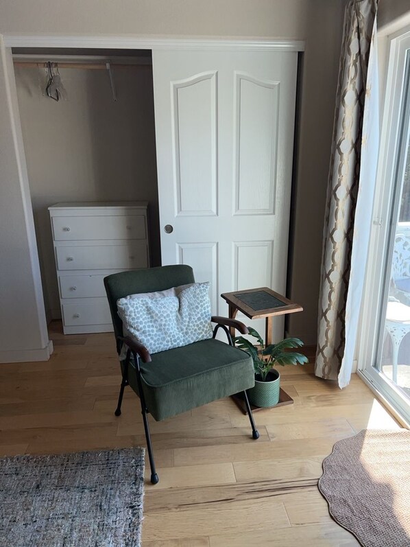 A room with a green armchair, a small table with a plant, a white dresser, and a door with a patterned curtain.