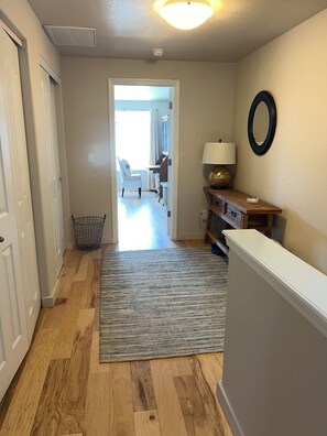 A hallway with a wooden floor, a grey rug, a lamp, a mirror, and a door leading to another room.