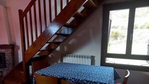 Una habitació amb una escala de fusta, una taula amb un mantell estampat, dues cadires, un radiador i una finestra amb vistes.