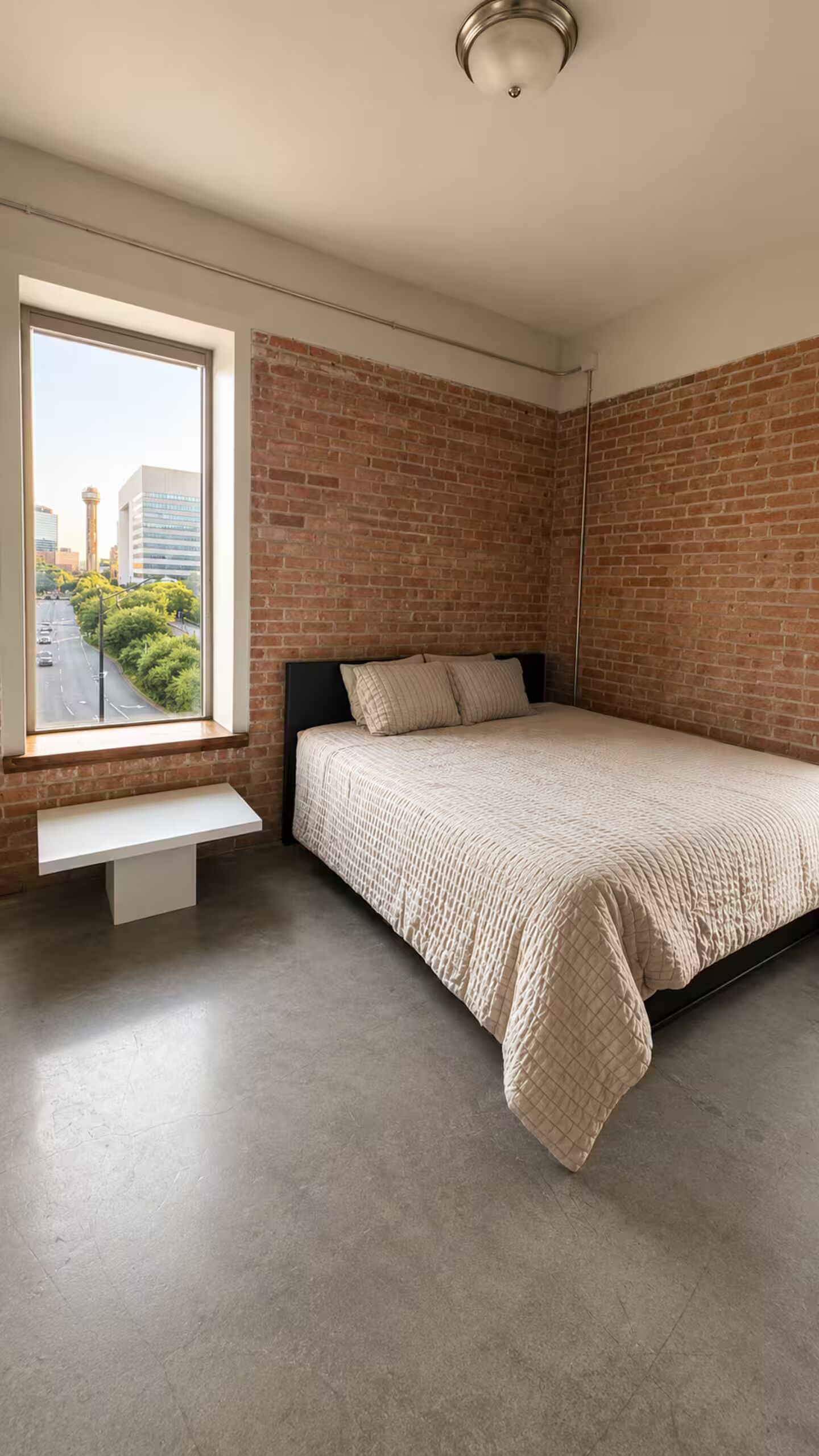 Une chambre avec un mur en briques, une fenêtre donnant sur des immeubles et de la verdure, un lit recouvert d’une courtepointe et un petit banc placé sous la fenêtre.