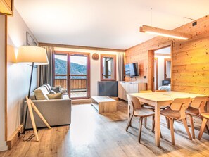 A modern room with a sofa, a dining table with chairs, a TV, and large windows with a mountain view. - Les Hauts de Vaujany by Interhome (Vaujany)