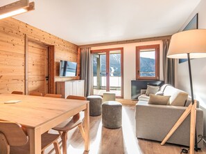 Ein Holzinnenraum mit Esstisch, Stühlen, einer Couch, Poufs, einem Fernseher und großen Fenstern mit Blick auf Berge.