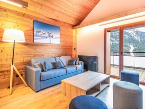 Ein gemütliches Zimmer mit Holzbalkendecke, einer Couch, einem Fernseher, einer Lampe und Blick auf Berge durch eine Glastür.