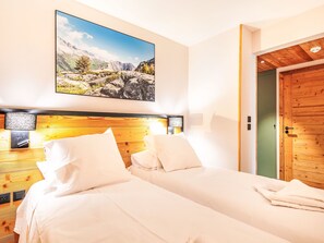 Ein Hotelzimmer mit einem Holzkopfteil, einem großen Landschaftsbild, zwei Betten mit weißen Leinentüchern und einer Holztür.